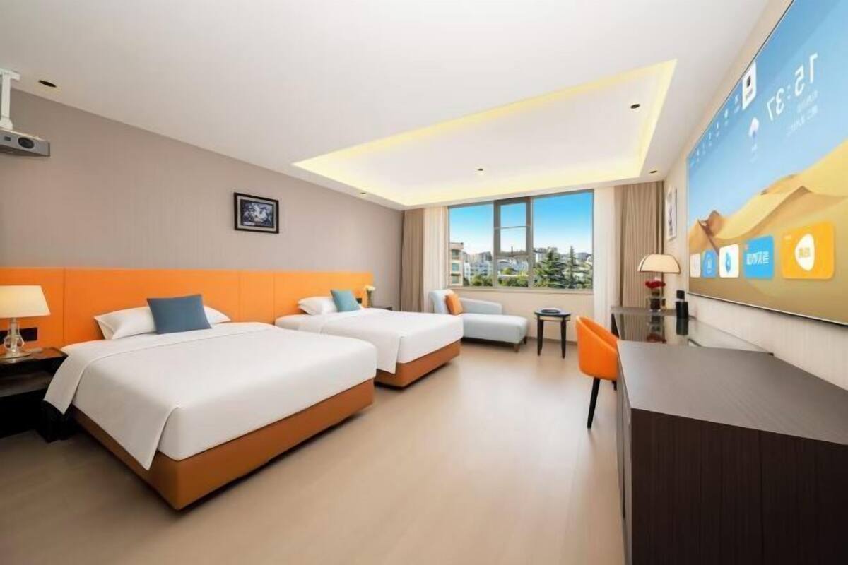 A hotel room with two beds, a desk, a chair, a window with a city view, and a large digital screen displaying information.