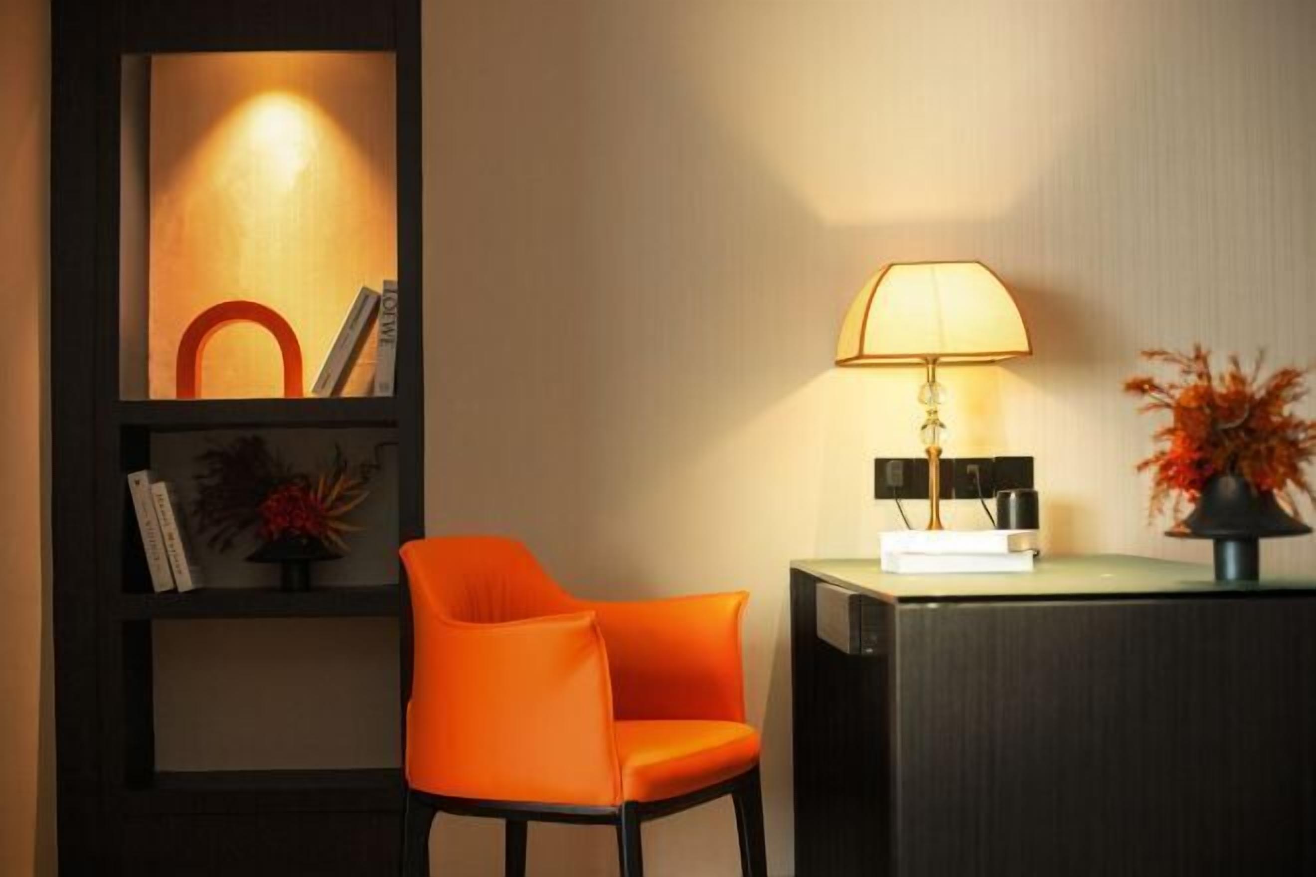 Una habitación moderna con un escritorio de madera oscura, una silla naranja brillante, una lámpara y una estantería con objetos decorativos.