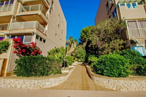 A pathway flanked by stone walls and lush greenery, leading to a multi-story residential building with balconies and windows.