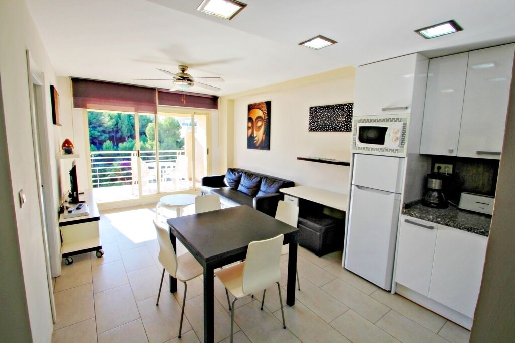 A modern kitchen with white cabinets, a microwave, and a coffee maker. There is a dining area with a table and chairs, and a sofa.