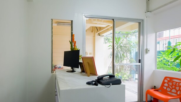 Un comptoir d’accueil équipé d’un écran d’ordinateur, d’un téléphone et d’une petite plante. Une porte-fenêtre coulissante donne accès à un espace extérieur verdoyant.