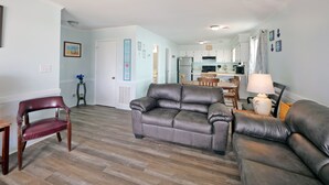 A spacious living room with a grey leather sofa, a wooden dining table, and a kitchen in the background. - "SeaQuest East" -2nd Row Pet Friendly Duplex, 4 Bedrooms / 3 Bathrooms (Holden Beach)