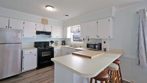 A modern kitchen with white cabinets, stainless steel appliances, a central island with stools, and a window providing a view. - "SeaQuest East" -2nd Row Pet Friendly Duplex, 4 Bedrooms / 3 Bathrooms (Holden Beach)