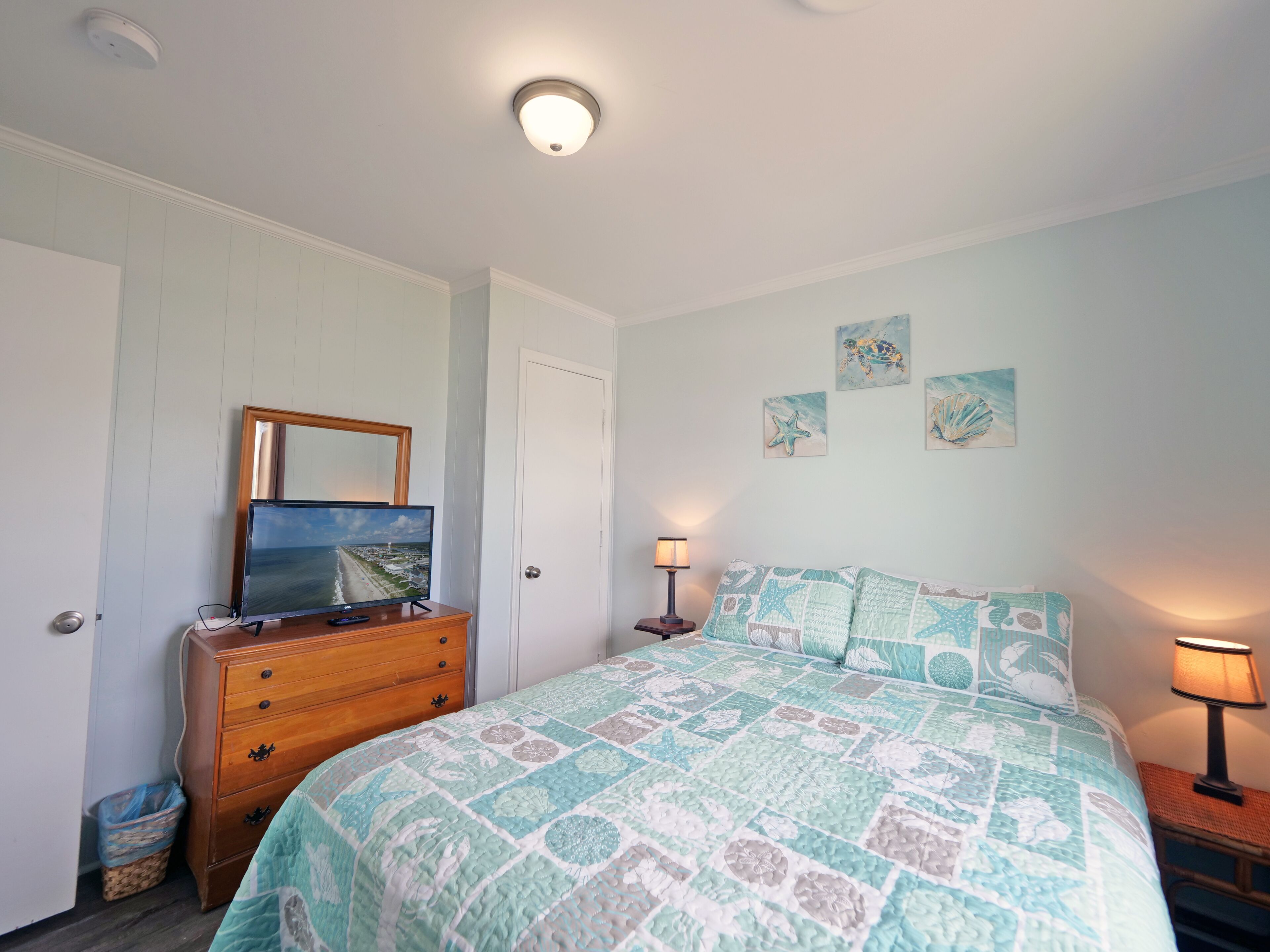 Une chambre à coucher avec un lit, une commode et un téléviseur posé sur la commode. On y trouve également des œuvres d’art murales et un luminaire au plafond.