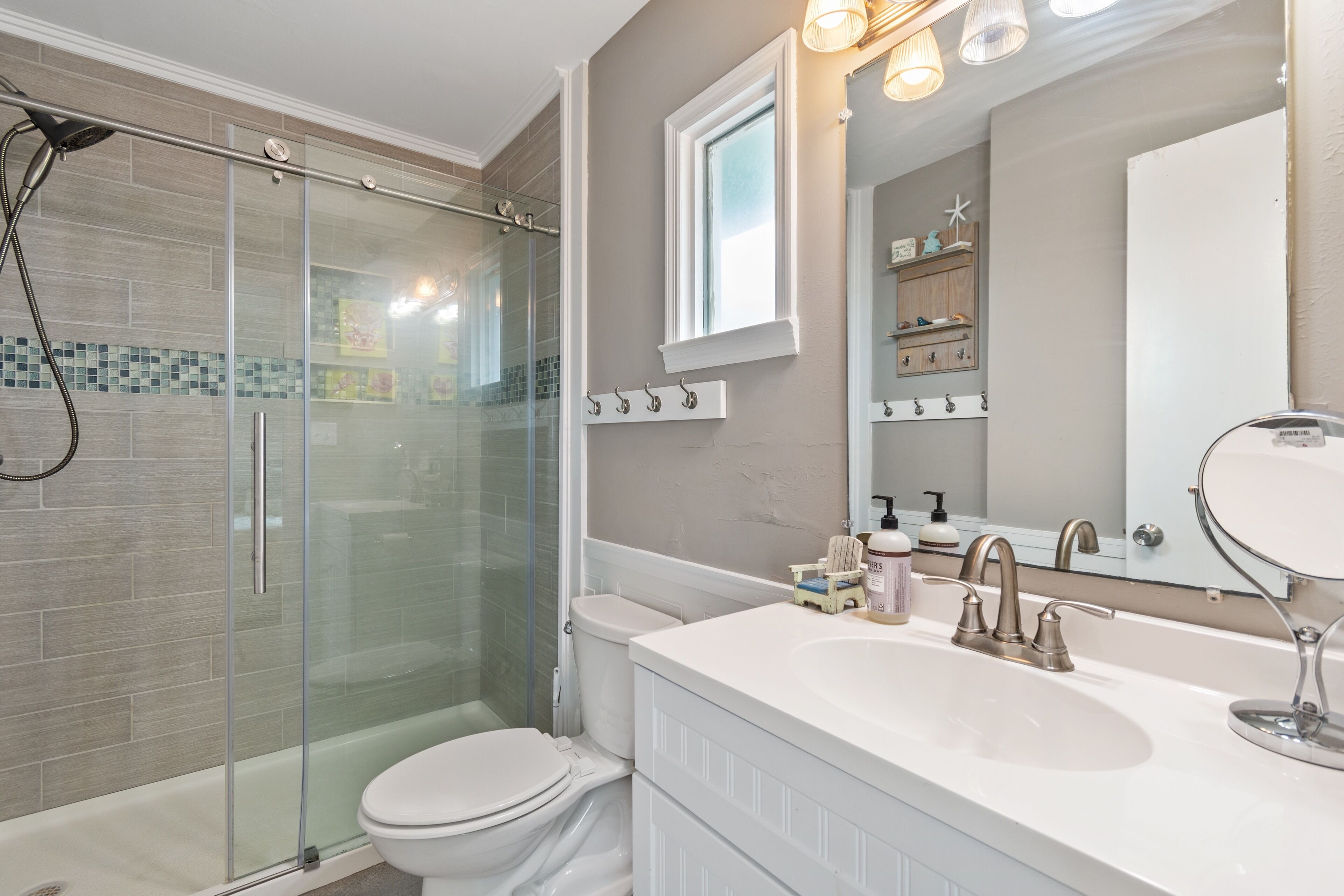 A modern bathroom with a glass-enclosed shower, a white toilet, a vanity with a large mirror, and a window above the mirror.