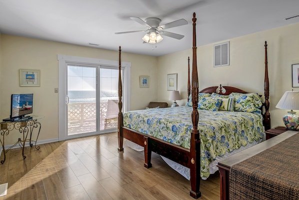 A four-poster bed with a floral bedspread, a ceiling fan, and a view of the ocean through a sliding glass door.