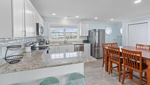 A modern kitchen with white cabinets, granite countertops, stainless steel appliances, and a dining area with wooden chairs and stools. - "Mocean Granted" - Oceanfront Home, 5 Bedrooms / 4 Bathrooms (Holden Beach)