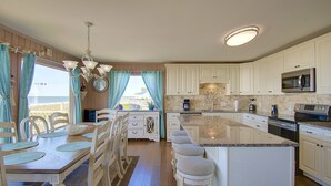 A spacious kitchen with white cabinetry, a central island with stools, a large window with curtains, and a chandelier. - "Goin' Coastal" - Oceanfront Home, 4 Bedrooms / 4.5 Bathrooms (Holden Beach)