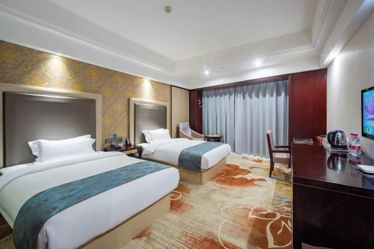 A hotel room with two beds, a desk, a chair, a TV, and a large window with curtains.