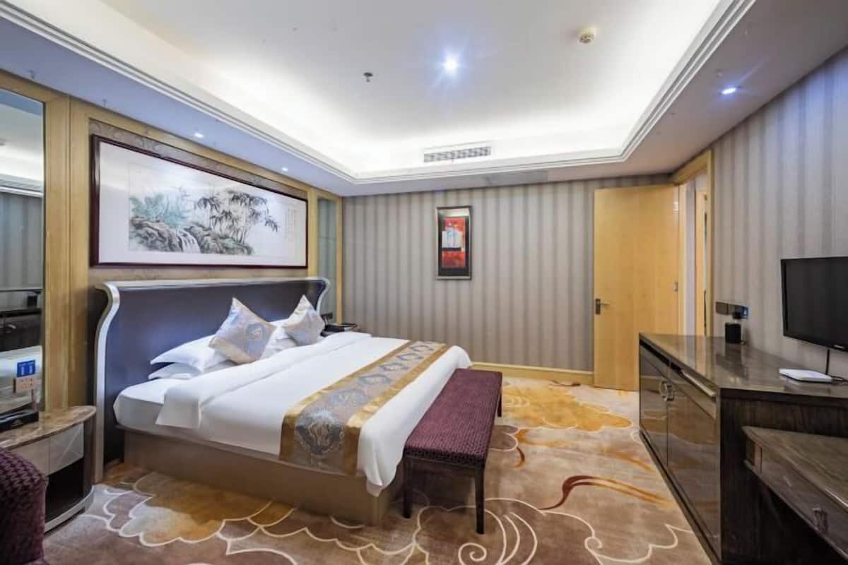A hotel room with a bed, a bench, a TV, a desk, and a framed picture on the wall.