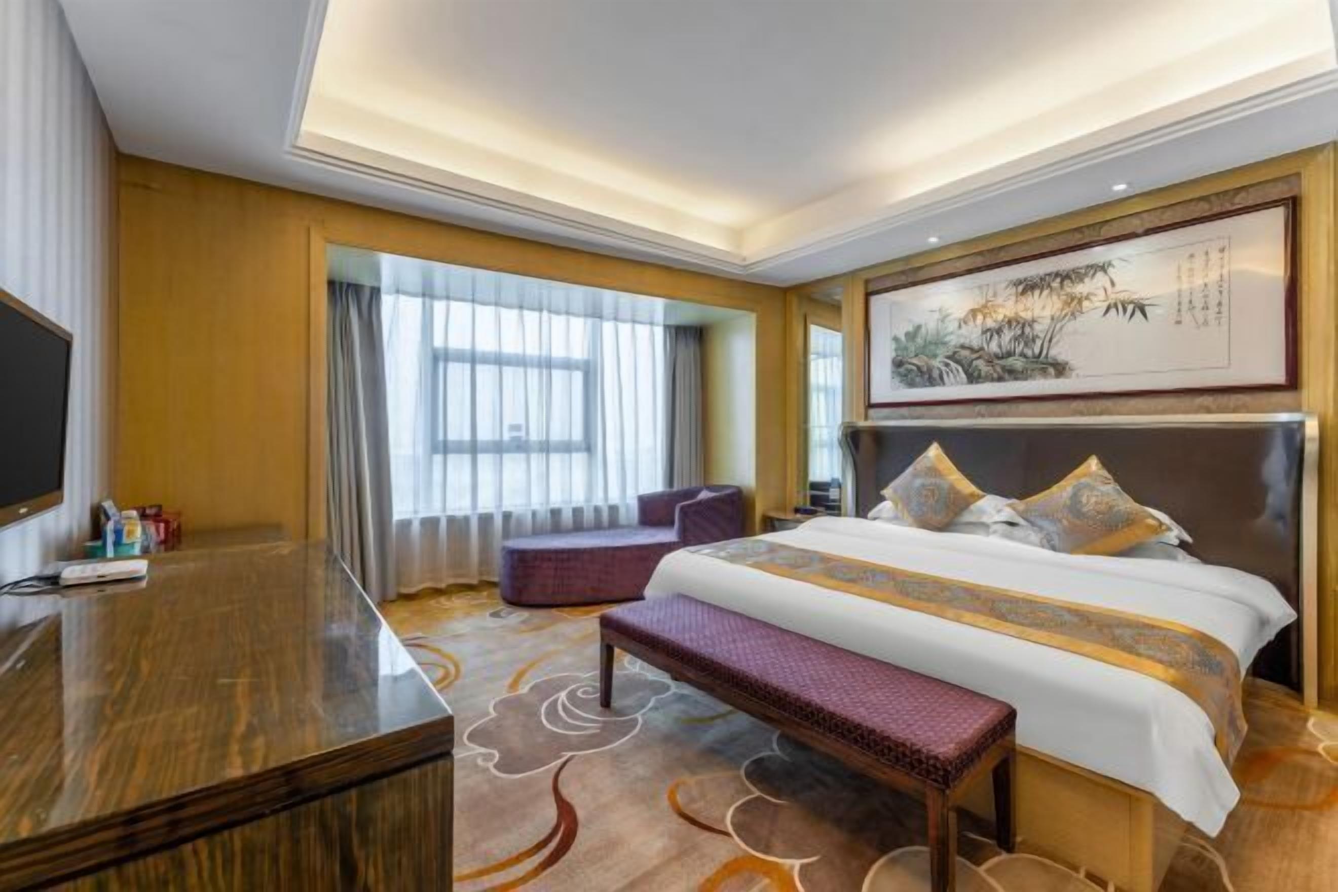 A hotel room with a large bed, a desk, a bench, and a television.
