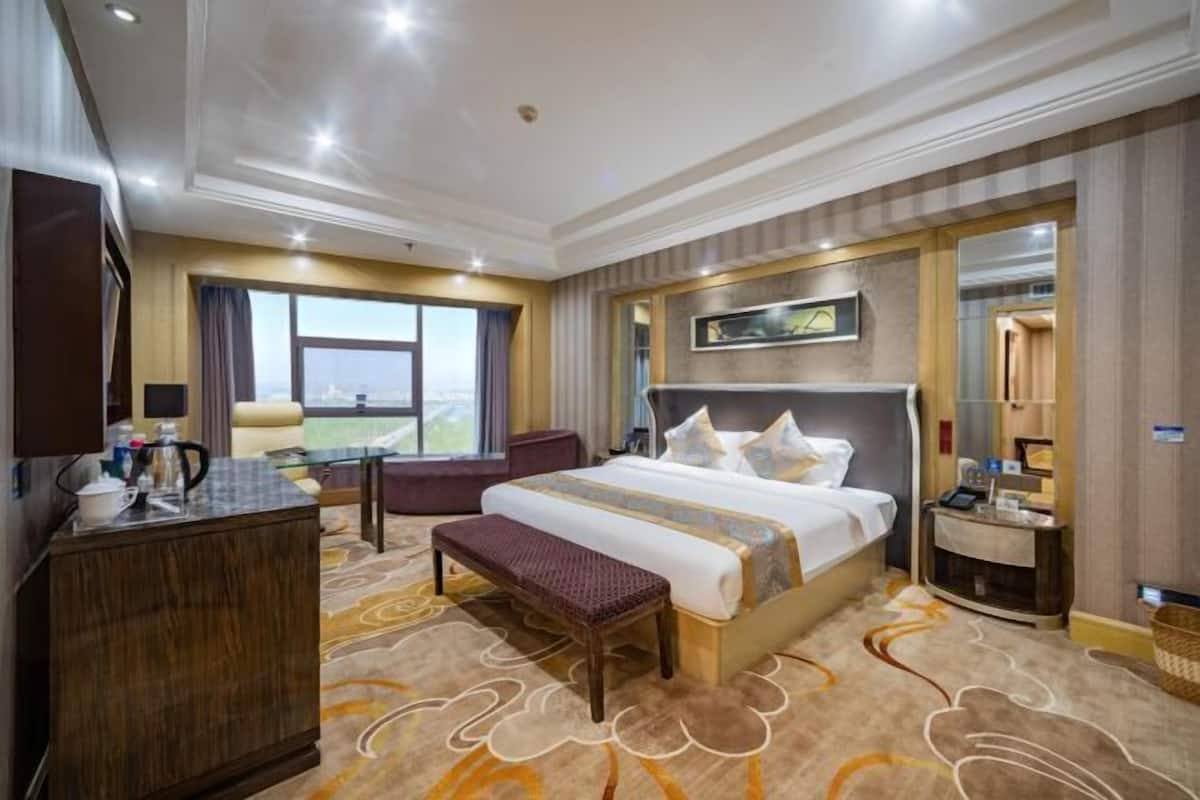 A hotel room with a large bed, a desk, a chair, a bench, and a view of the city.