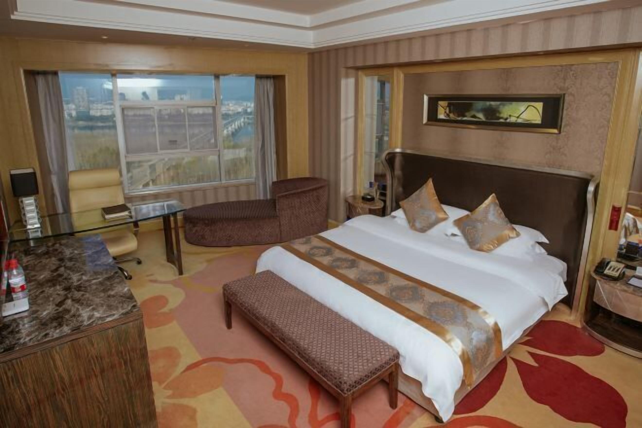 A hotel room with a large bed, a desk, a chair, and a view of a cityscape through the window.