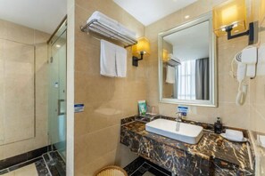 A hotel bathroom with a glass shower enclosure, a marble vanity with a sink, a large mirror, and a hairdryer mounted on the wall.