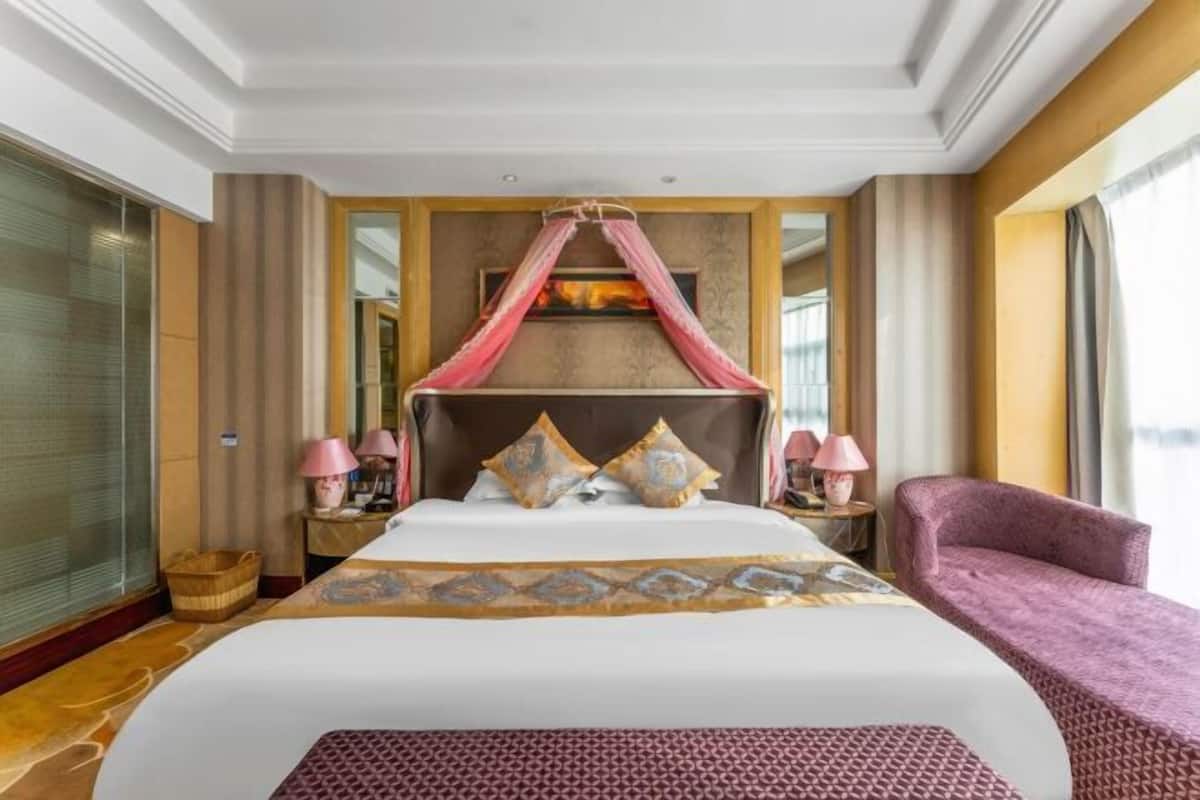 A hotel room with a large bed, a canopy, two bedside tables with lamps, a purple armchair, and a large mirror on the wall.
