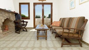 Una sala de estar acogedora con una chimenea de piedra, una mesa de madera, un sofá a rayas y dos amplios ventanales con vistas al exterior.