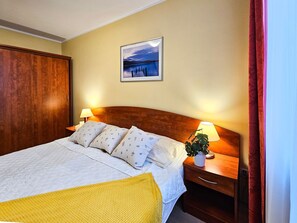 Habitación de hotel con cabecera de madera, cama con ropa de cama blanca y almohadas estampadas, mesita de noche con lámpara y un cuadro enmarcado en la pared.