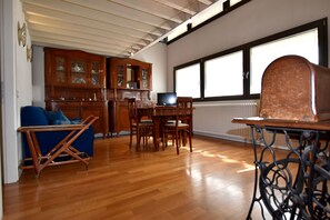 A spacious living room with wooden flooring, a blue sofa, a dining table with chairs, a television, and a large window with a view.