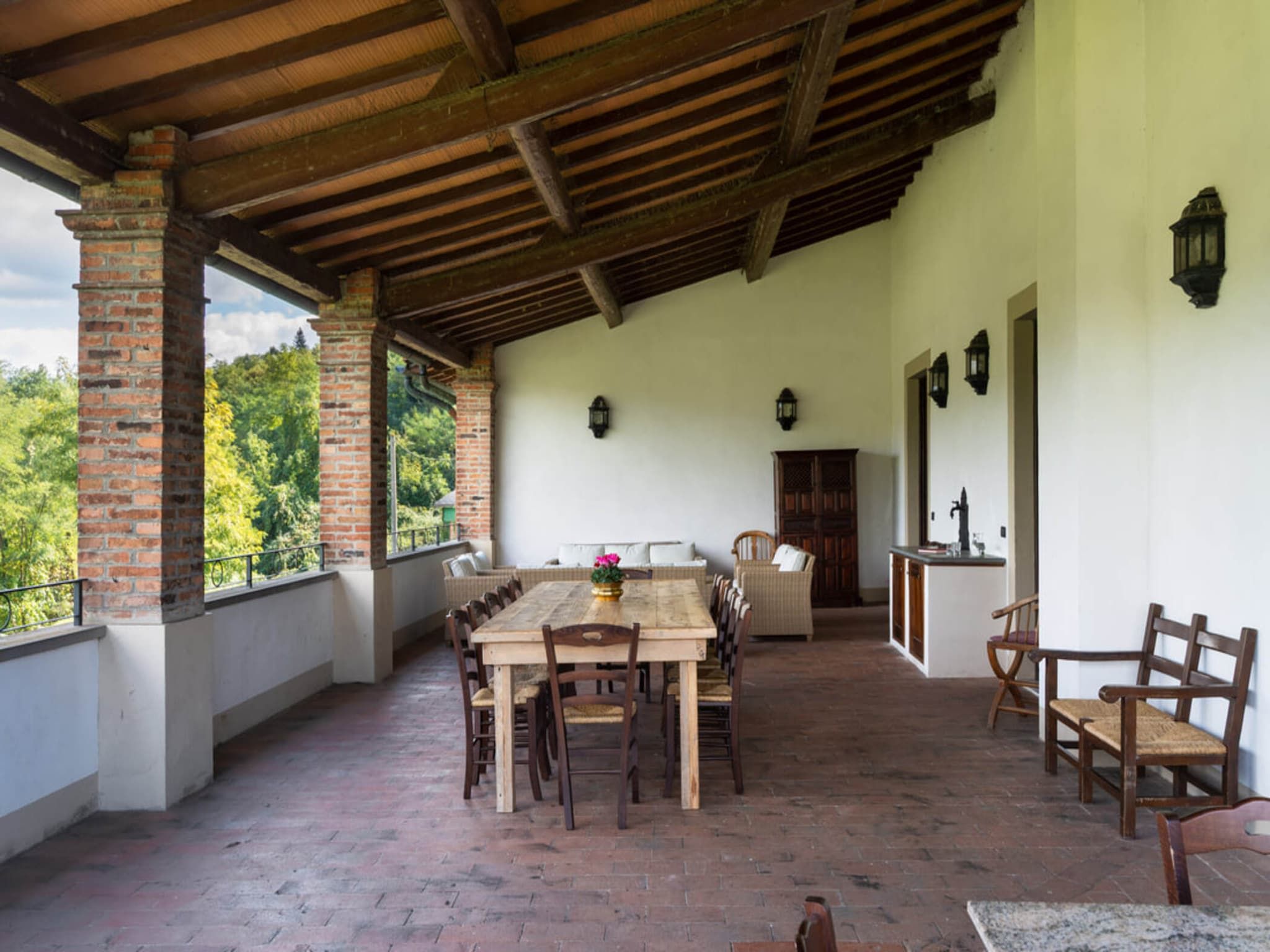 A covered patio with a wooden table and chairs, a brick chimney, and a view of greenery.