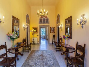 A hallway with a chandelier, two side tables with lamps, chairs, a rug, and a door leading to another room.