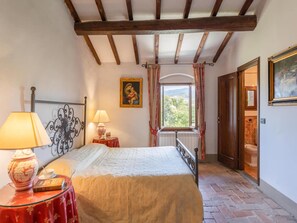 A bedroom with a bed, a nightstand, a lamp, a painting, a window with curtains, and a door leading to a bathroom.