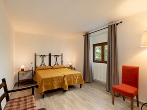 A bedroom with a double bed, a chair, a table with a lamp, and a window with curtains.
