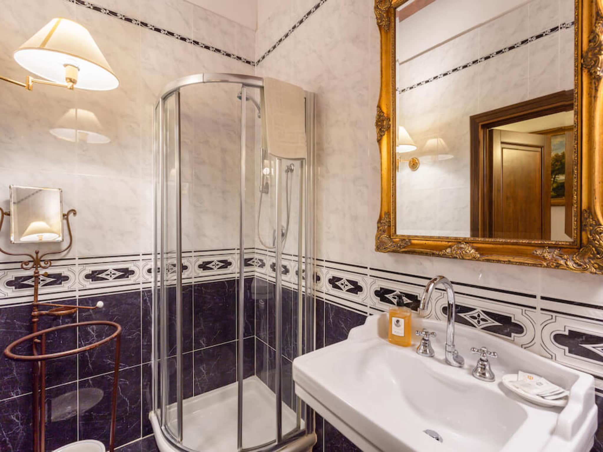A bathroom with a glass-enclosed shower, a white sink with a gold soap dispenser, and a large ornate mirror.
