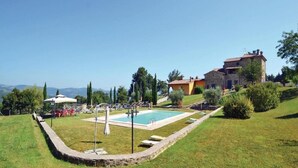 A pool area with a stone border, a covered seating area, and a building with a red roof.