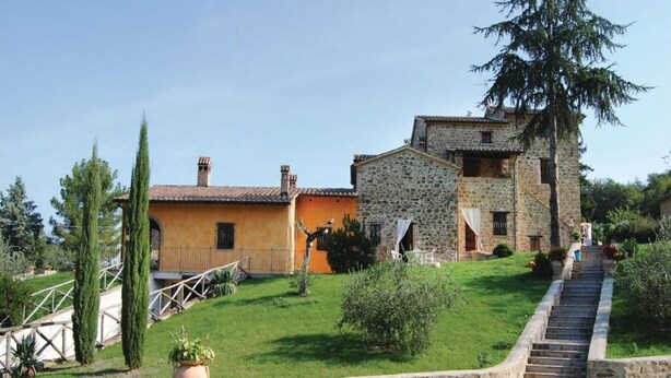 A stone building with a staircase leading up to a door, surrounded by greenery and a clear sky.