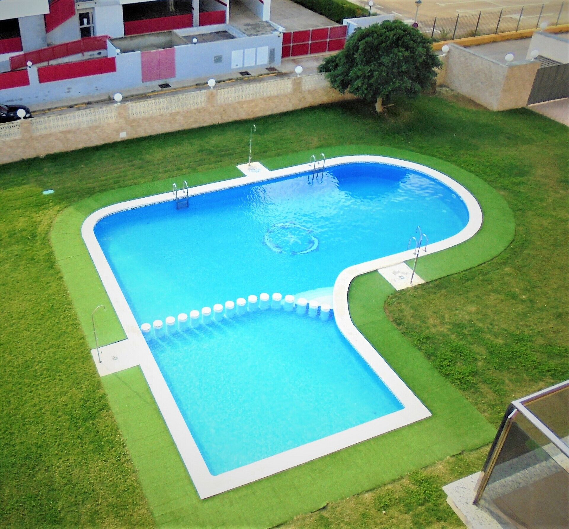 Ein Pool mit einem ungewöhnlichen L-förmigen Design, umgeben von gepflegtem Rasen und einer niedrigen Mauer.