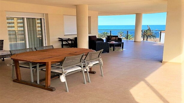 Un balcón amplio con una mesa y sillas de madera, vista al océano y un edificio al fondo.