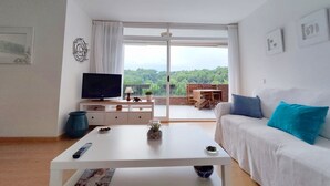 A modern living room with a television, a white coffee table, a sofa with blue cushions, and a balcony with a view of greenery. - Apto Residencial en Tamariu with Mountain View, Pool, Wi-Fi & Garden (Tamariu)