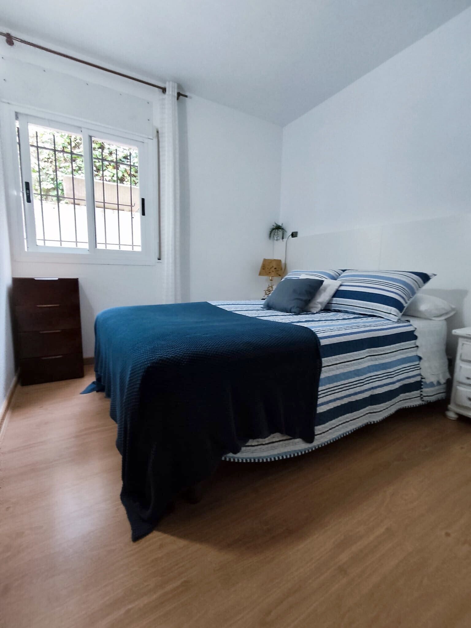 Un dormitorio con cama, mesita de noche, lámpara, ventana con rejas y piso de madera.