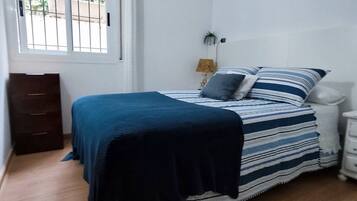 Un dormitorio con cama, mesita de noche, lámpara, ventana con rejas y piso de madera.