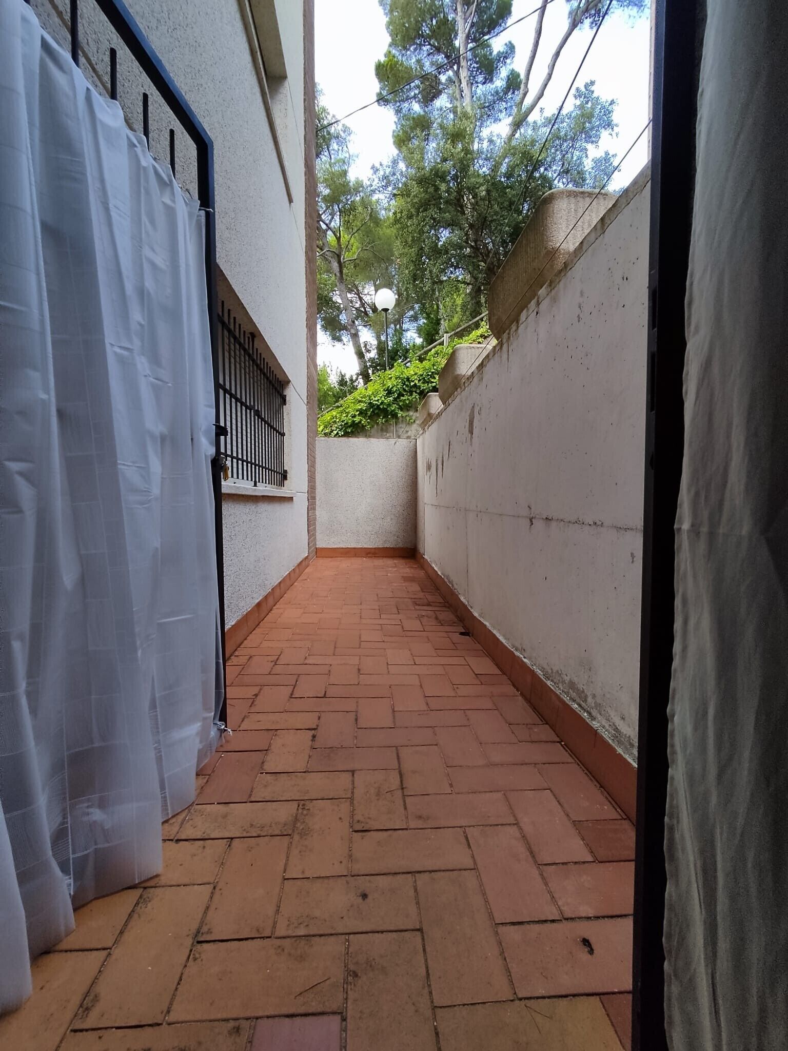 Un estrecho sendero de ladrillo que conduce a una entrada protegida por una cortina blanca, un portón de metal y un muro blanco con un pequeño ventanal.