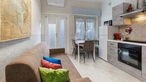 Eine moderne Küche mit Backofen, ein Essbereich mit Tisch und Stühlen sowie ein Wohnzimmer mit Sofa und bunten Kissen.