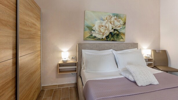 Ein ordentlich bezogenes Bett mit weißen Leinen und Kissen, ein Nachttisch mit Lampe, ein Blumenbild an der Wand und ein Holzschrank.