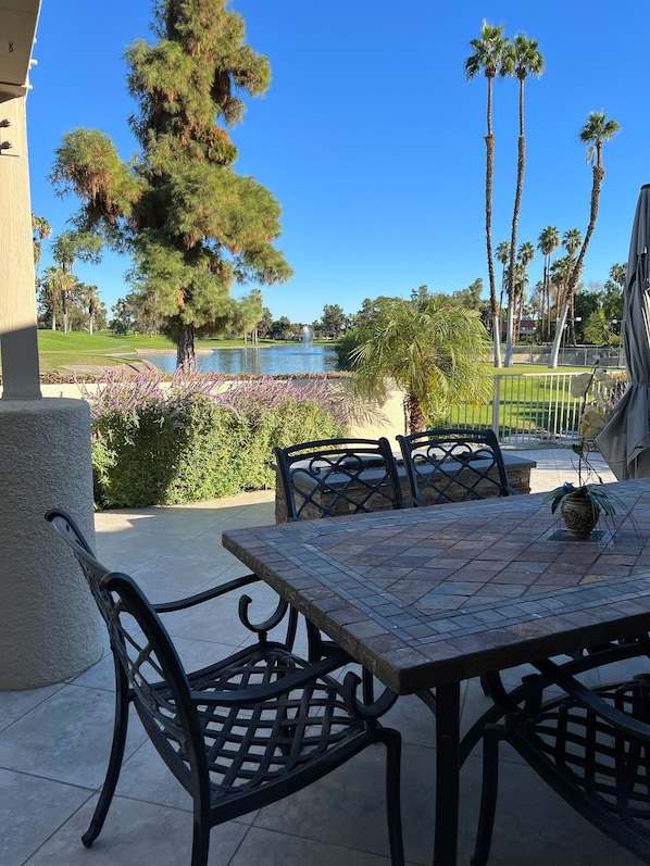 Une terrasse extérieure avec une table et des chaises, entourée d’un jardin aménagé avec différentes plantes, et offrant une vue sur un terrain de golf et un plan d’eau.