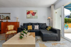 A living room with a sofa, a wooden chair, a coffee table with flowers, a painting on the wall, and a sliding glass door leading to an outdoor area.