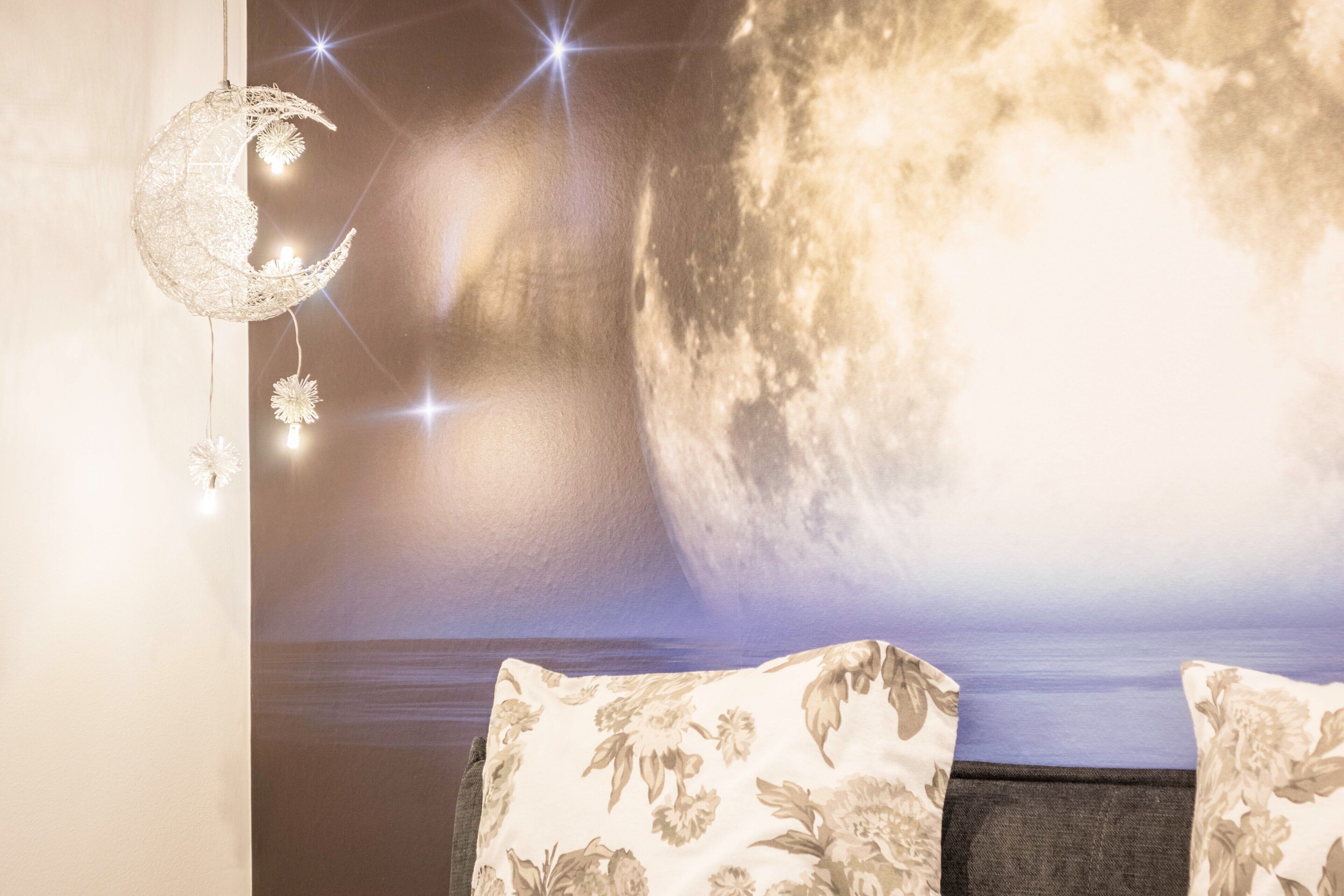 Une pièce dont un mur est décoré d’une fresque représentant la lune et les étoiles, avec une décoration en forme de lune suspendue et deux coussins à motifs floraux sur un lit.