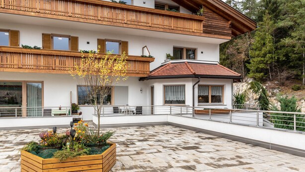 A two-story building with a balcony, a wooden planter with flowers, and a tiled patio area. - Apartment "Pramstaller Kron" with Mountain View, Wi-Fi, Terrace & Garden (St. Lorenzen)