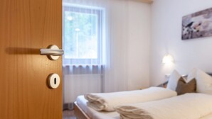 A hotel room with a wooden door, a bed with white bedding, a window with sheer curtains, and a wall-mounted light fixture. - Apartment "Pramstaller Kron" with Mountain View, Wi-Fi, Terrace & Garden (St. Lorenzen)