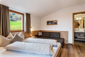 Une chambre d’hôtel avec une grande fenêtre, un canapé, un lit avec une literie à carreaux, un tableau au mur et une salle de bain avec un miroir et un lavabo.