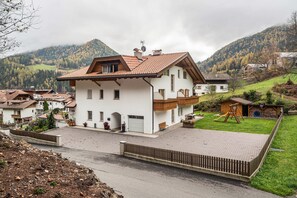 Une maison à deux étages, dotée d’un toit en pente, d’une cour clôturée et offrant une vue sur un paysage montagneux.