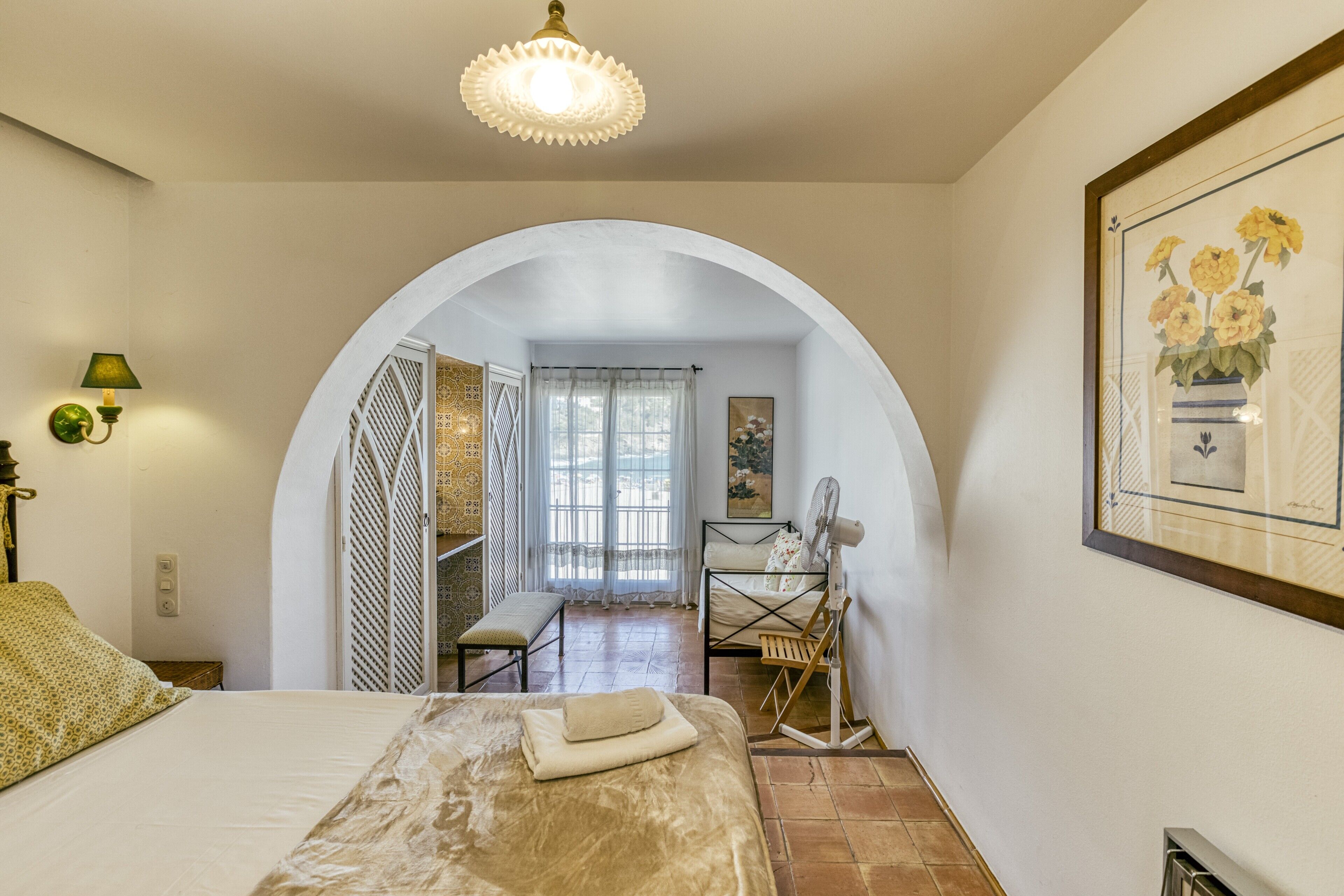 A bedroom with a bed, a fan, a bench, a door leading to another room, and a framed picture on the wall.