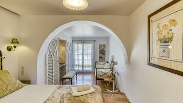 A bedroom with a bed, a fan, a bench, a door leading to another room, and a framed picture on the wall.