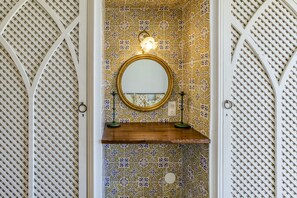 A bathroom with a patterned tile wall, a round mirror, and a wooden shelf.