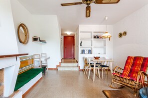 Ein Wohnzimmer mit einer Couch, einem Esstisch mit Stühlen, einem Schaukelstuhl aus Weidengeflecht, einem Deckenventilator und einem Bücherregal.