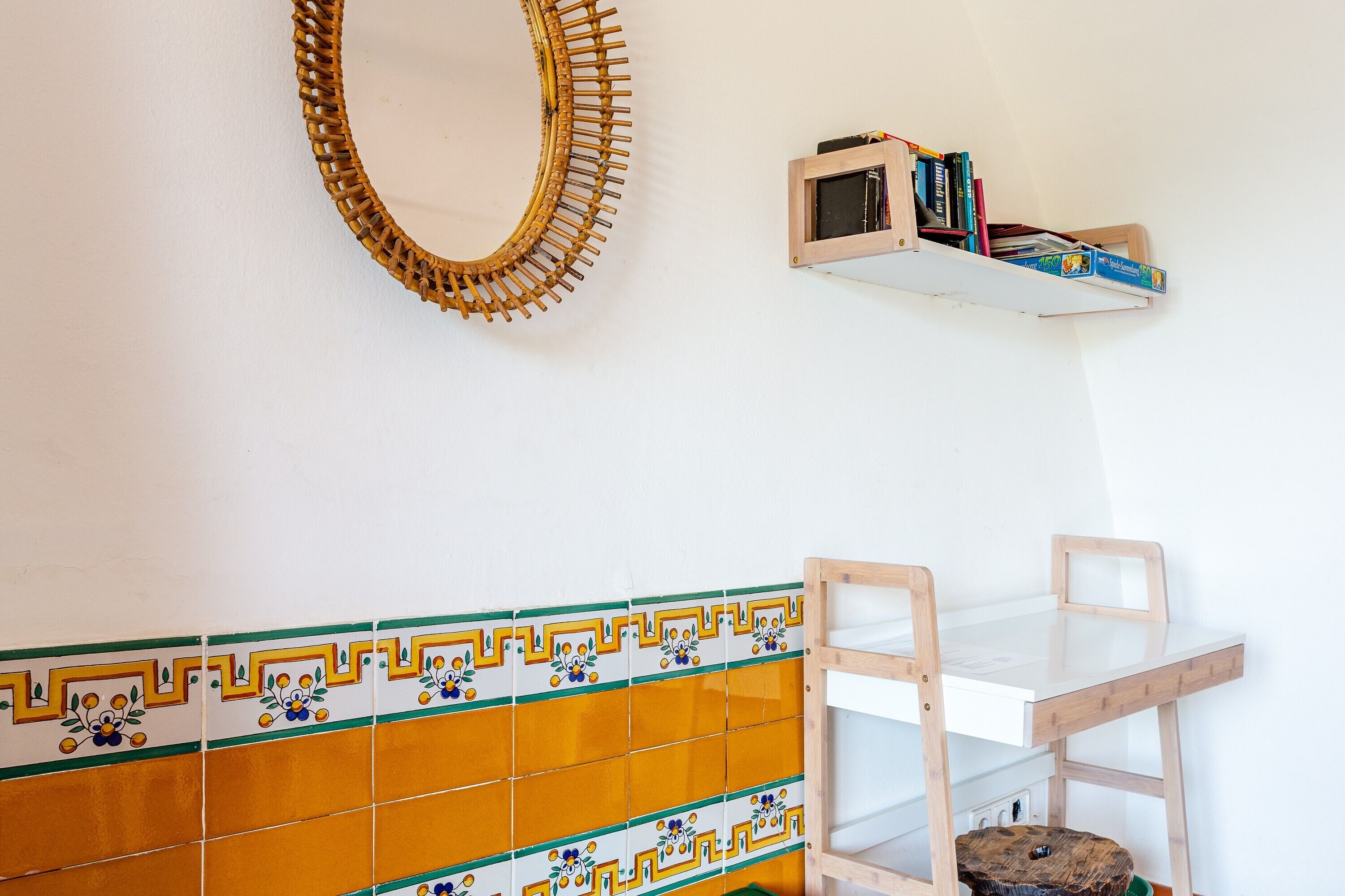 Een kamer met een witte muur, een spiegel met houten lijst, een plank met boeken, een betegelde achterwand en een houten stoel.