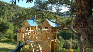 A wooden playhouse with a blue roof, a slide, and a swing set in a grassy area with trees and hills in the background. - Exclusive Villa with Pool, Sea View, and Wi-Fi (Tossa De Mar)